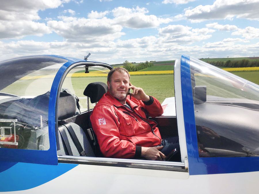 Fritz Madl in roter Fliegerjacke sitzt telefonierend im geöffneten Cockpit eines blau-weißen Ultraleichtflugzeugs