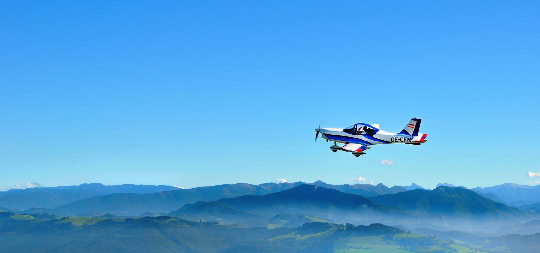 Weiß-blau-rotes Ultraleichtflugzeug mit der Kennung OE-CFM im Flug über den bewaldeten Hügeln des Mühlviertels bei strahlend blauem Himmel