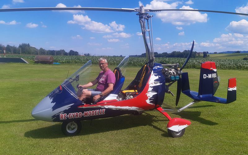 Blau-rot lackierter Tragschrauber mit Pilot im Cockpit auf einer grünen Wiese vor blauem Himmel